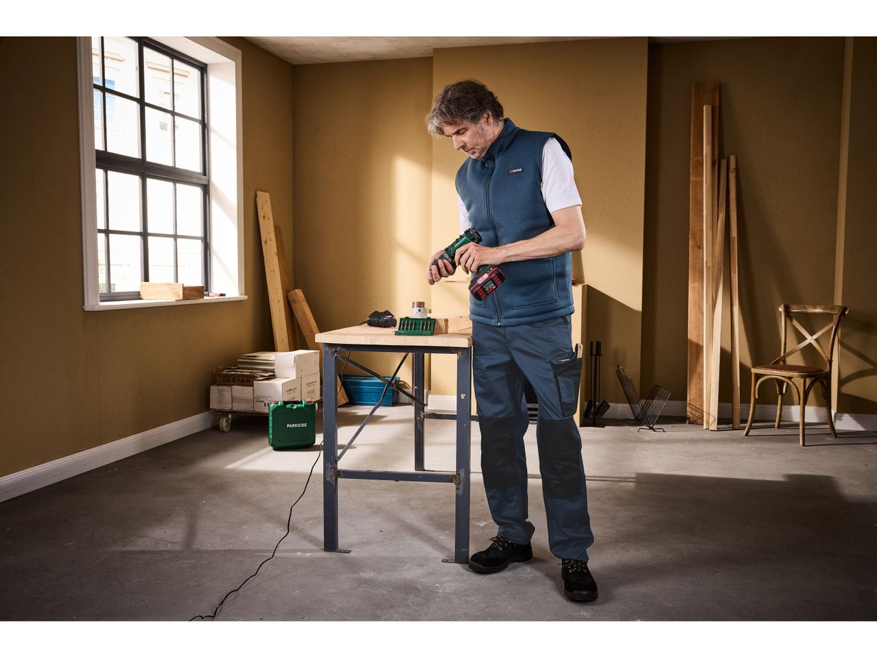 Un uomo in un gilet blu e pantaloni da lavoro tiene un trapano avvitatore accanto a un banco da lavoro.