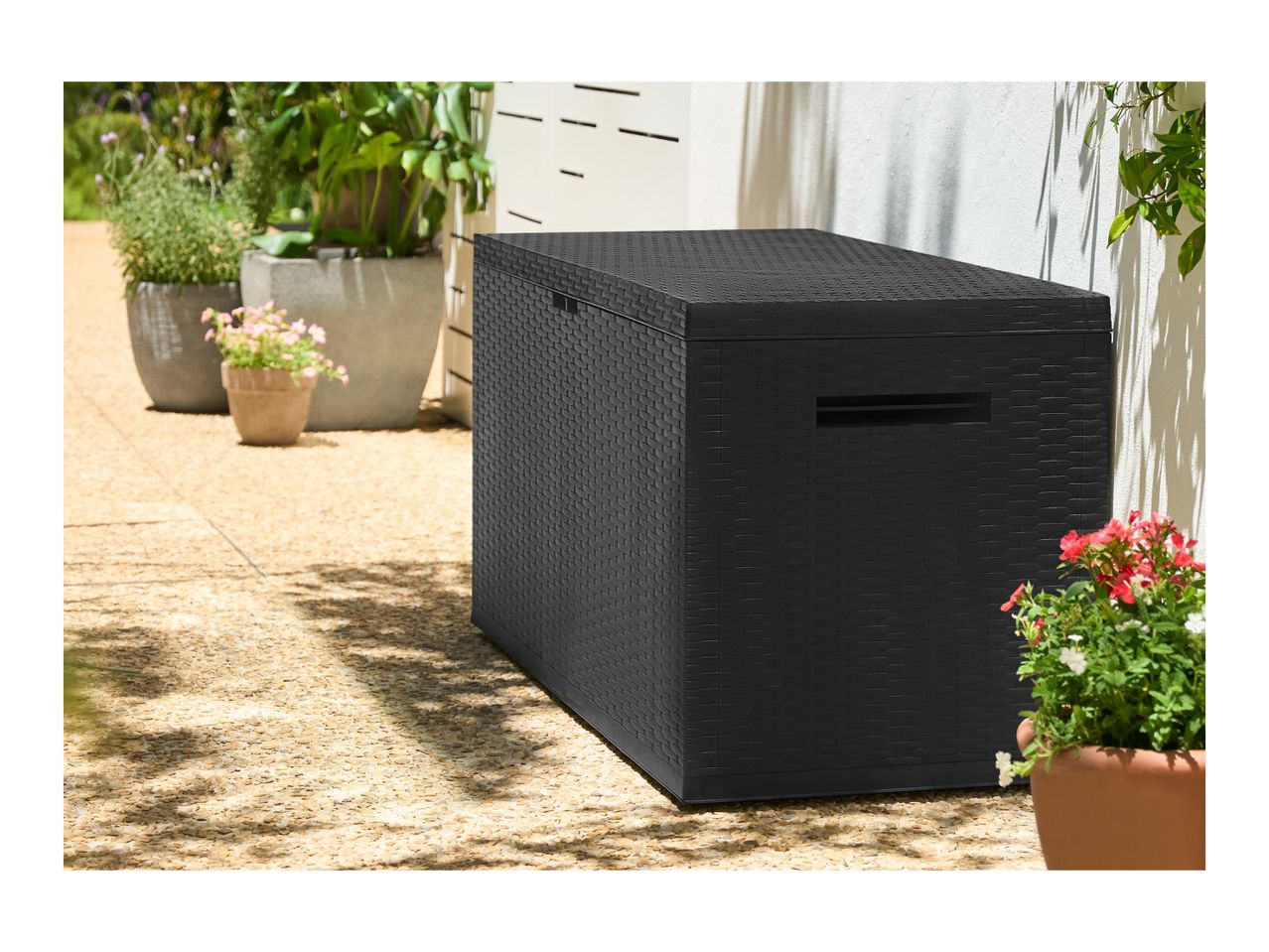 Black rattan-style garden storage box on a patio with potted plants.