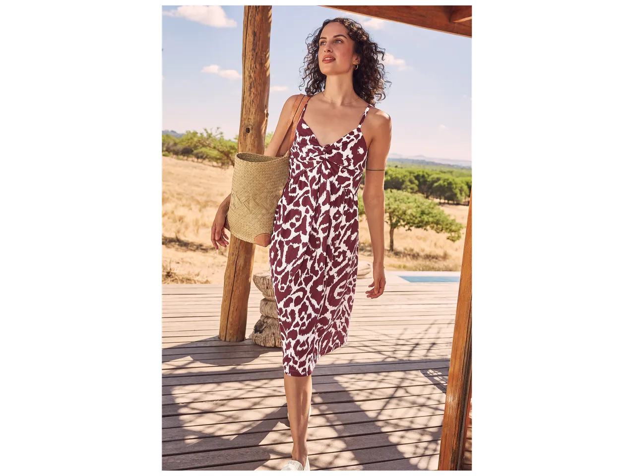 Une femme en robe d'été à motif animalier et sac de plage en paille sur une terrasse en bois.