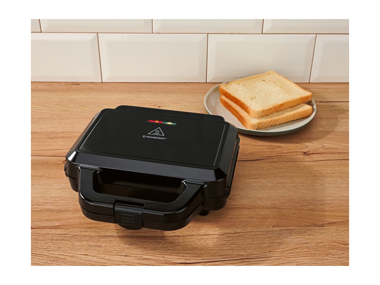 Black sandwich maker on a wooden surface with two slices of bread on a plate.