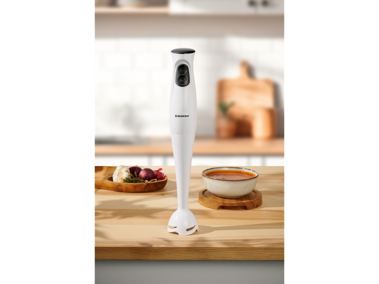A white SilverCrest immersion blender on a wooden counter with a bowl of soup and ingredients.