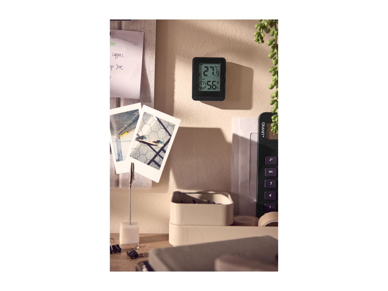 A digital thermometer displaying 27.9°C and 56% humidity, surrounded by office supplies.