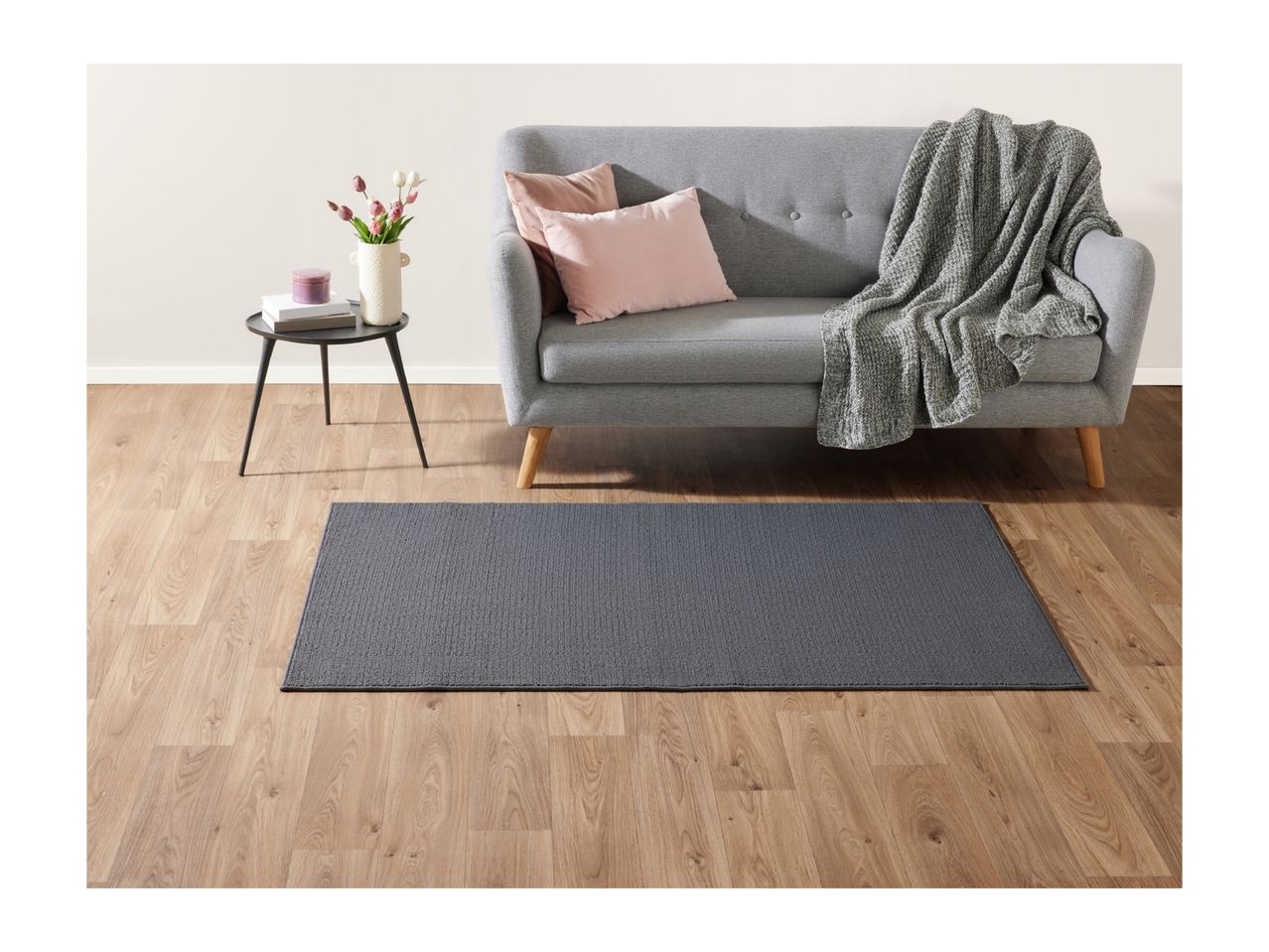 A gray sofa with pink pillows and a throw blanket, a side table with flowers, and a gray rug on a wooden floor.