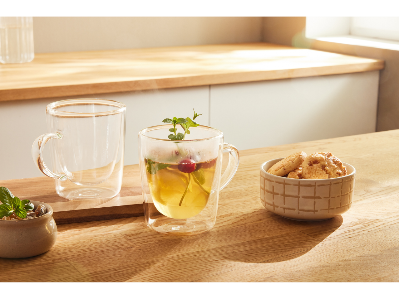 Divas stikla tējas krūzes un bļoda ar cepumiem uz koka galda.