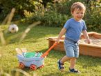 A happy toddler pulls a toy wagon with sand toys next to a sandbox in a sunny garden.