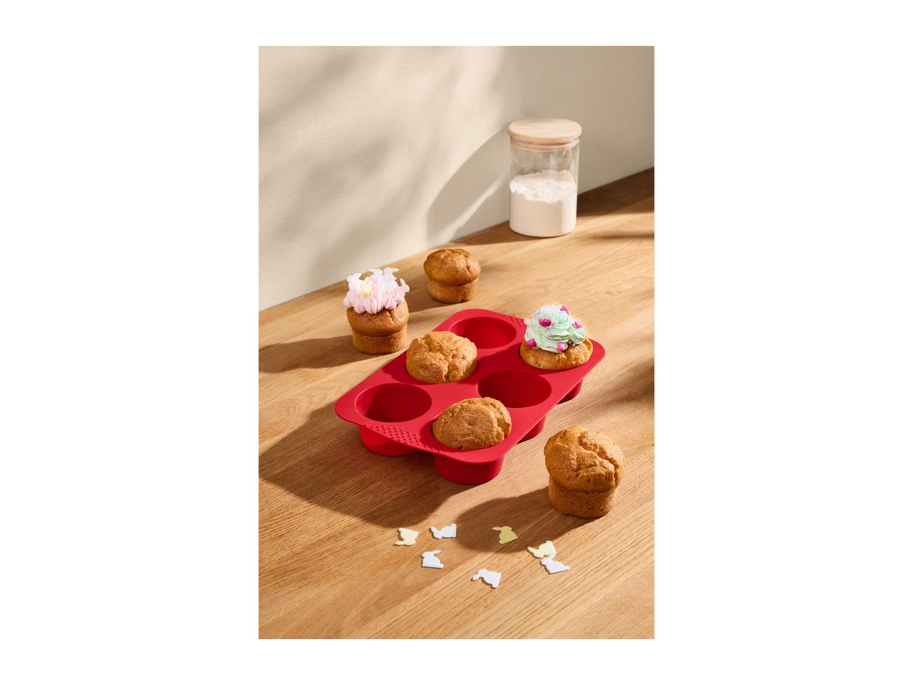Muffins in a red silicone baking tray, some decorated with frosting and sprinkles, on a wooden table.
