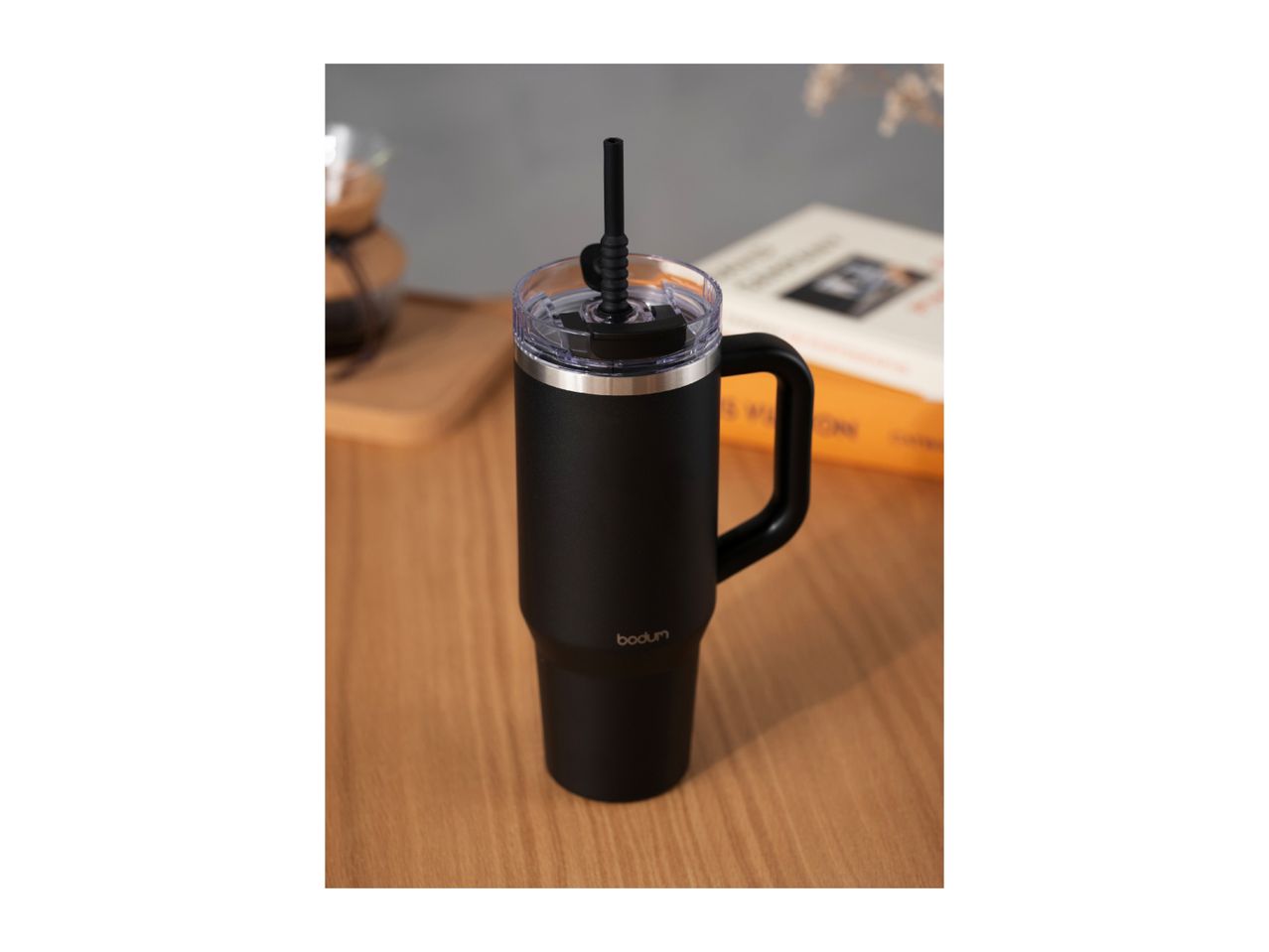 Black travel mug with a handle and straw on a wooden table, with a book and coffee maker in the background.