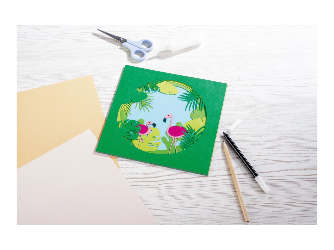 A tropical-themed paper craft with flamingos, surrounded by craft supplies like scissors, glue, and paper.