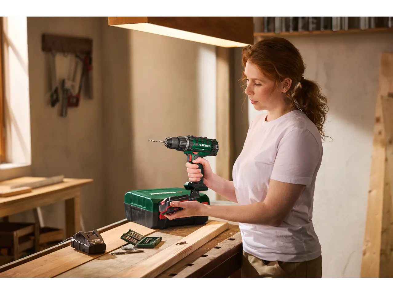 Woman holding a PARKSIDE® 20V Cordless Drill Driver, with a tool case and bits on a workbench.
