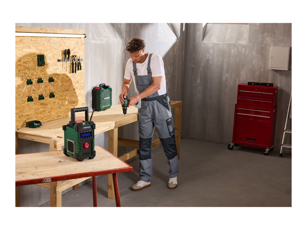 Man in overalls using a drill at a workbench with a portable radio and tool case.