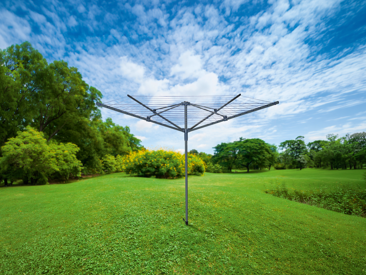 Περιστρεφόμενη απλώστρα ρούχων σε ένα καταπράσινο πάρκο κάτω από γαλανό ουρανό με λευκά σύννεφα.