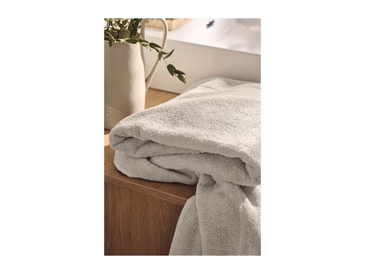 Folded light grey towels on a wooden surface next to a white vase with green foliage.