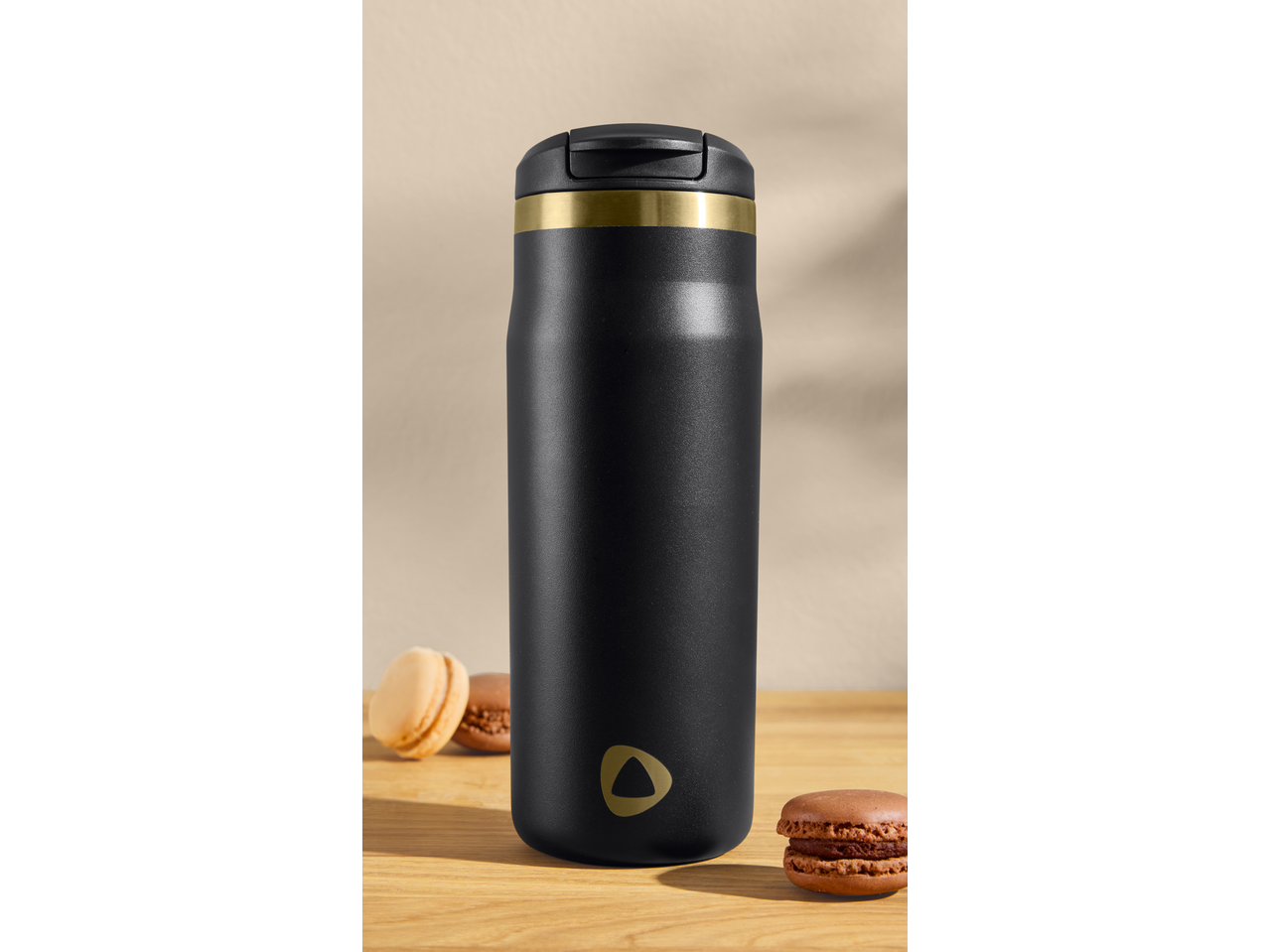 Black travel mug with a gold band and logo, next to two macarons on a wooden surface.