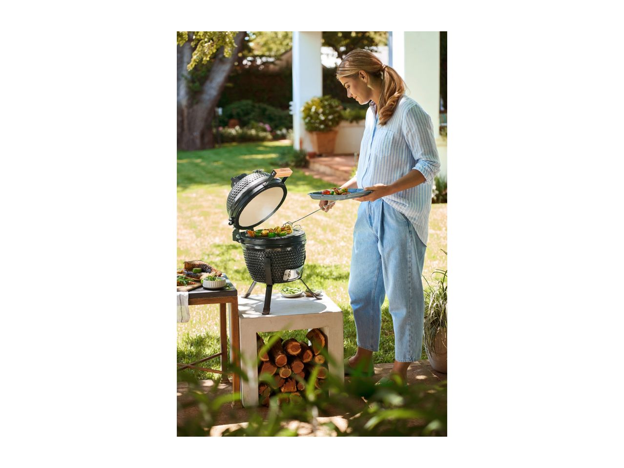 Woman grilling skewers on a portable ceramic barbecue in a garden setting.