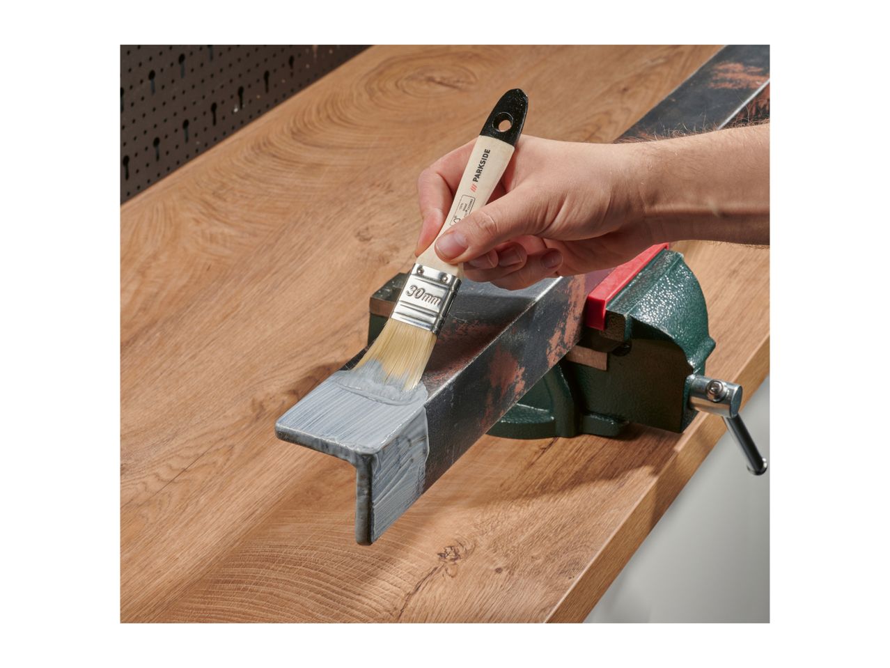 Hand painting a metal angle with a brush, secured in a vise on a wooden workbench.