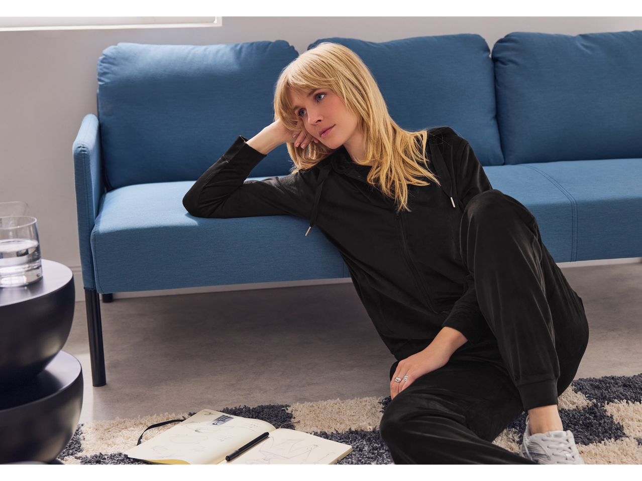 Γυναίκα με μαύρη βελούδινη φόρμα κάθεται στο πάτωμα δίπλα σε έναν μπλε καναπέ.