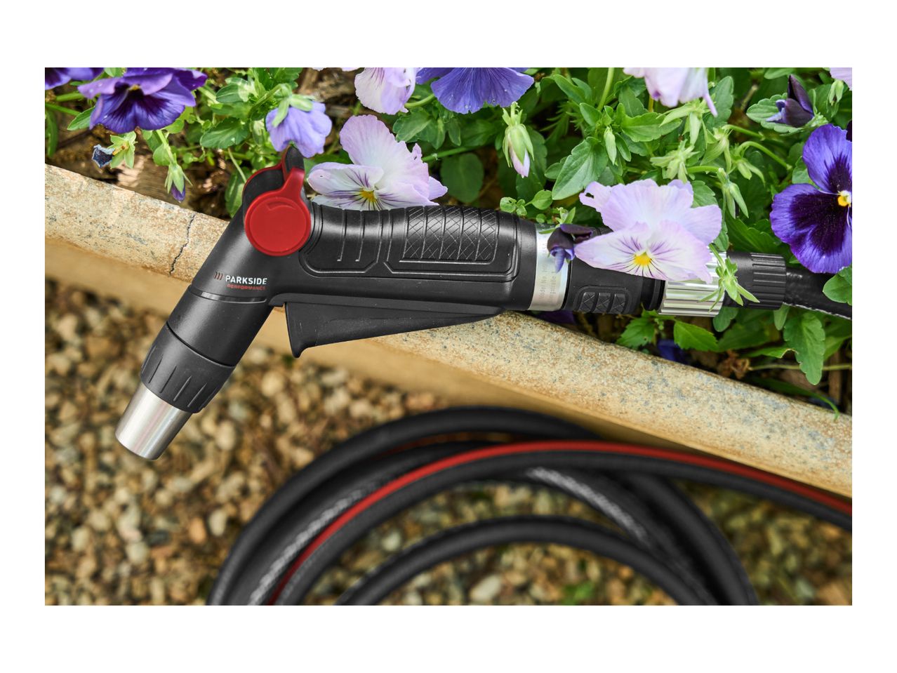 A black garden hose nozzle with a red trigger rests on a stone edge next to purple and white pansies.