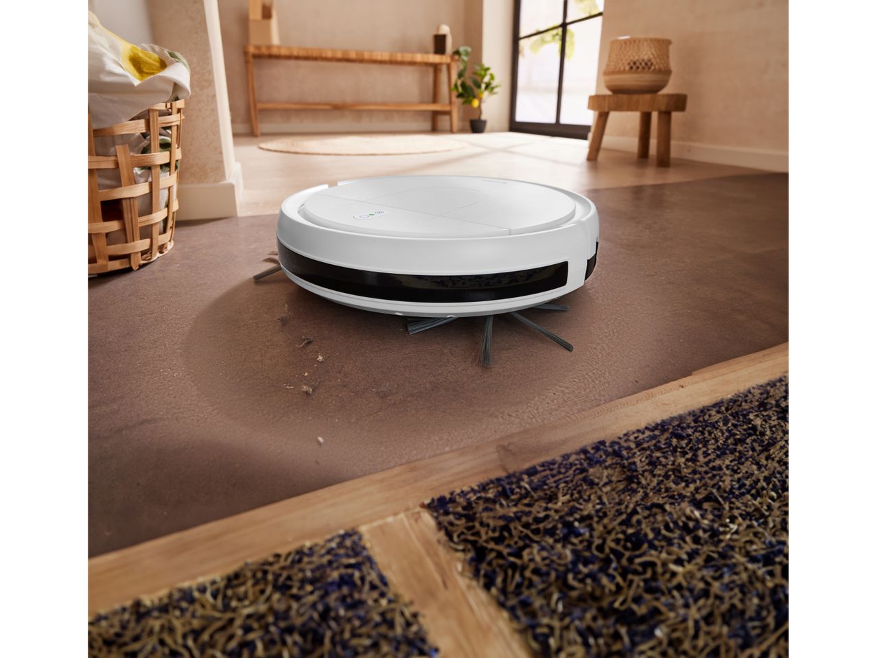 A white SILVERCREST® Robot Vacuum Cleaner cleans a brown floor in a modern home.