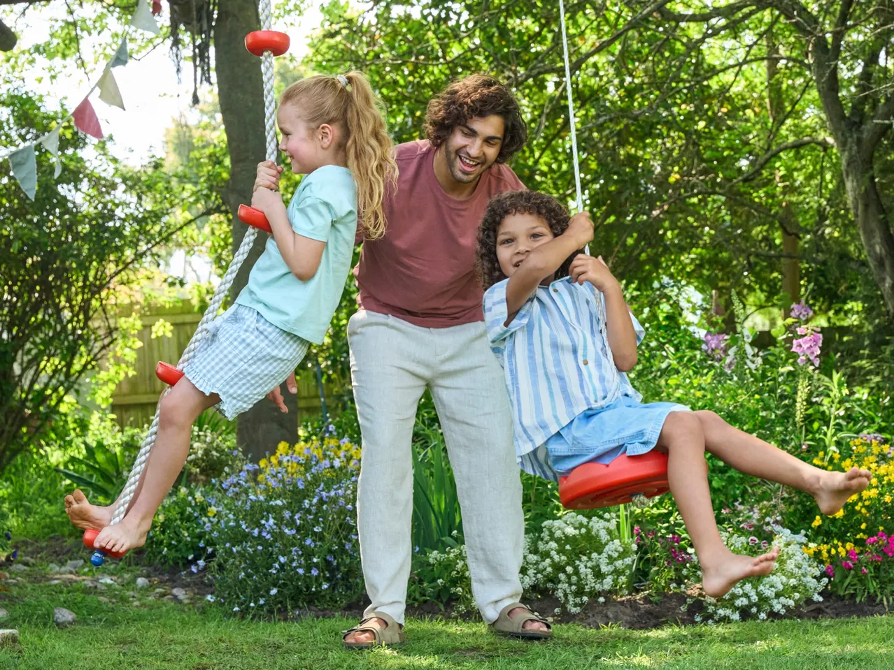 Father and two children playing on swings in a garden with colorful flowers and green trees.