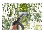 Hand holding a window cleaner, cleaning a soapy window with green foliage outside.