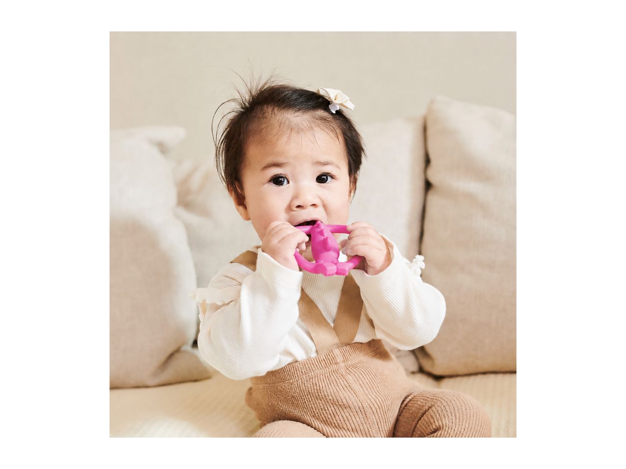 Baby in a white top and brown overalls, chewing on a pink teether.
