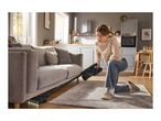 Woman vacuuming under a sofa with a cordless stick vacuum cleaner in a modern living room.