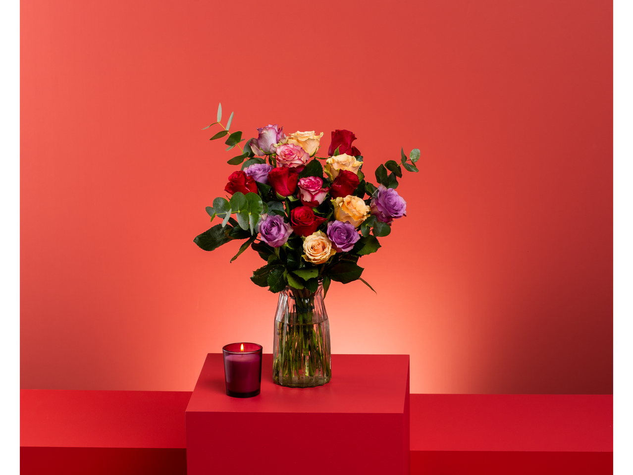 Colorful rose bouquet in a glass vase with a lit candle on red display blocks.