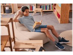 Un garçon lit un livre, assis sur un pouf, avec des écouteurs, dans une pièce avec des étagères.
