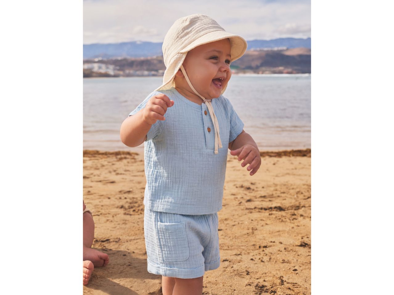 Happy baby in a light blue muslin outfit and sun hat, playing on a sandy beach.