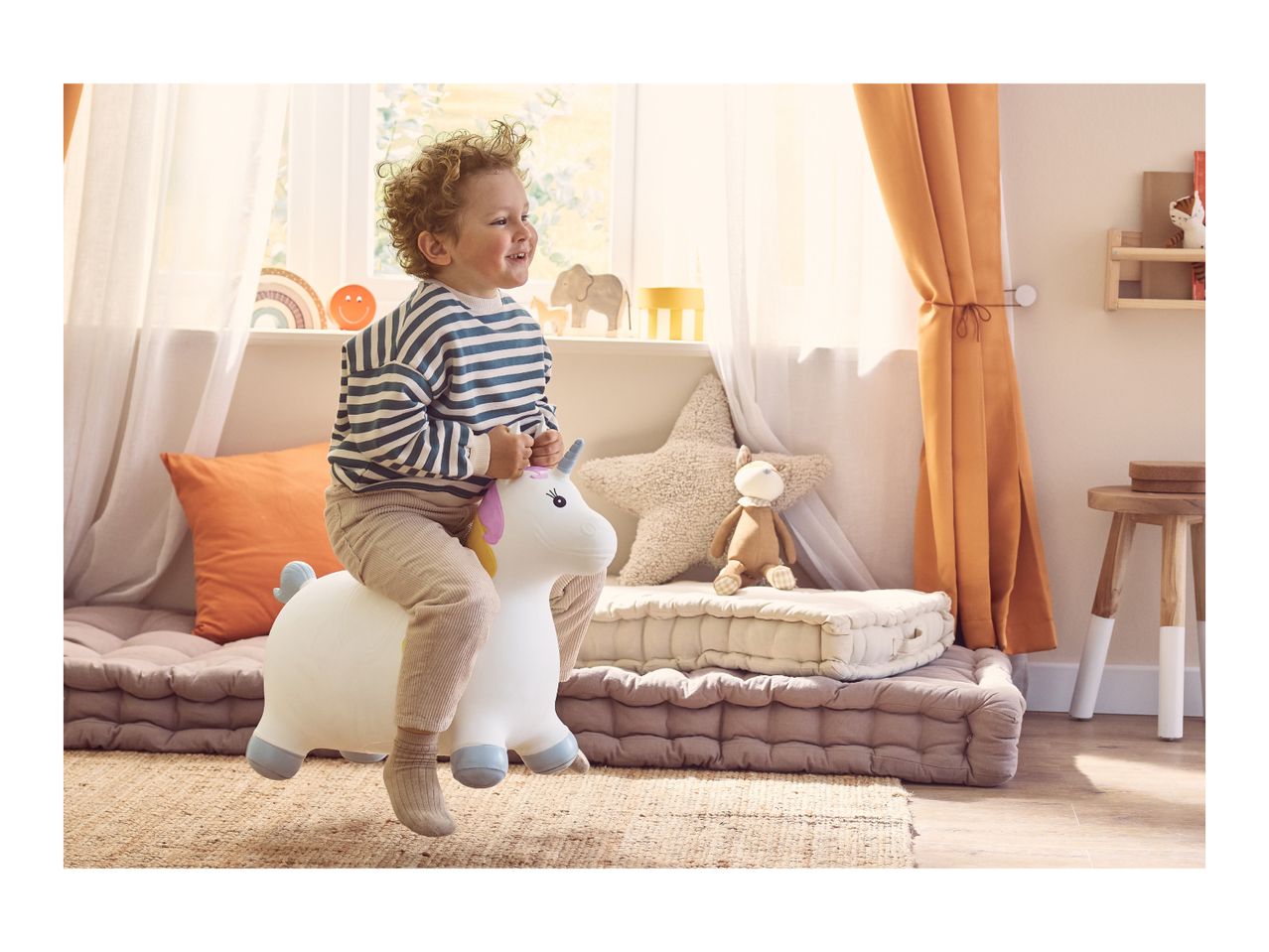 Happy child on a unicorn bouncy toy in a bright, cozy playroom with floor cushions.