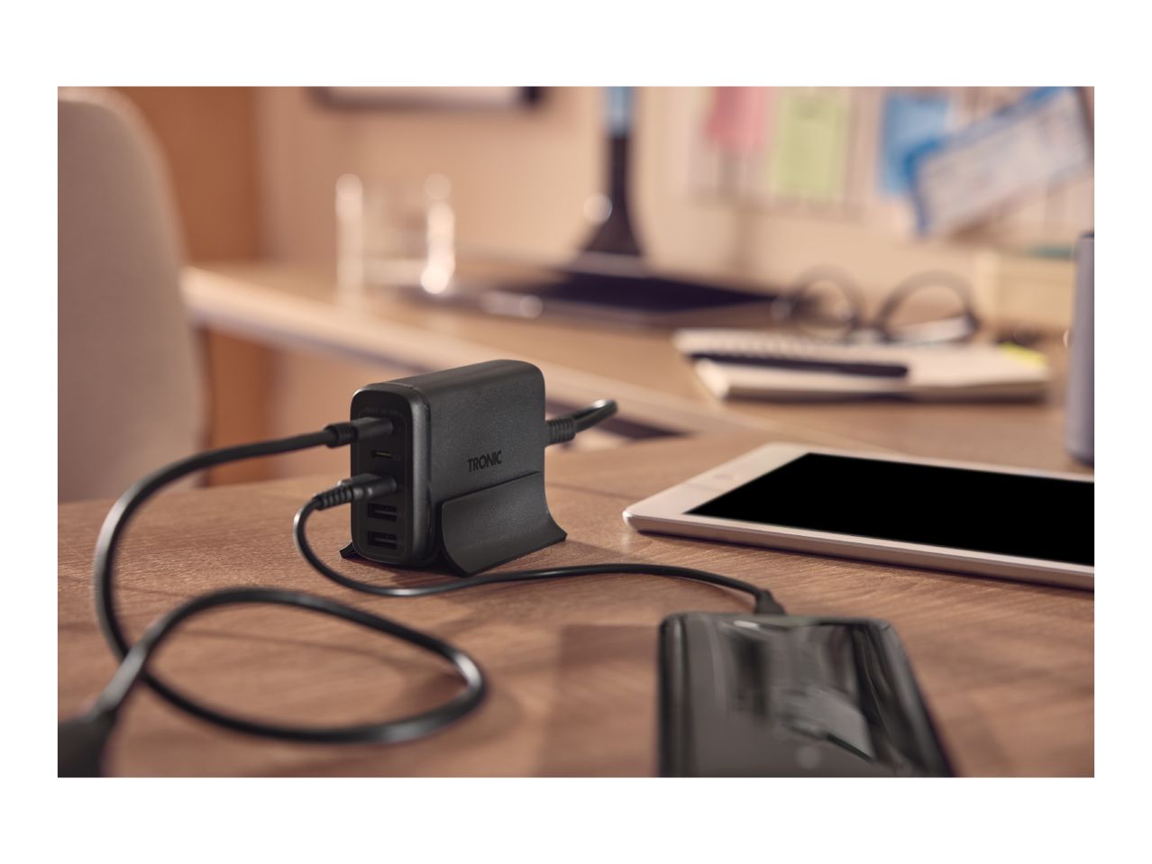 Black multi-port USB charger with TRONIC logo, charging a smartphone and tablet on a wooden desk.