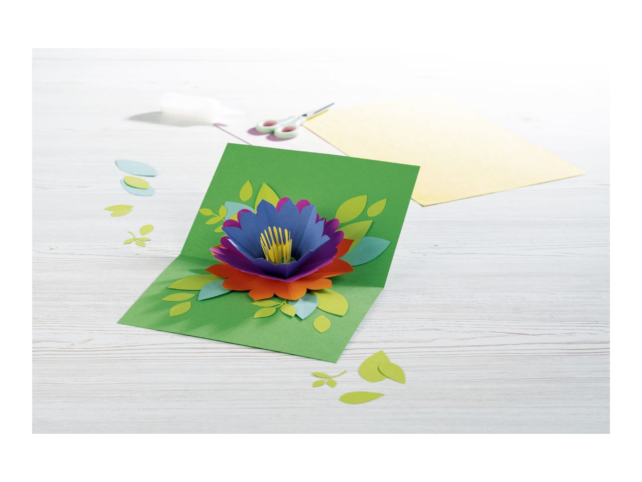 A vibrant, handmade pop-up flower card with crafting supplies on a white wooden table.