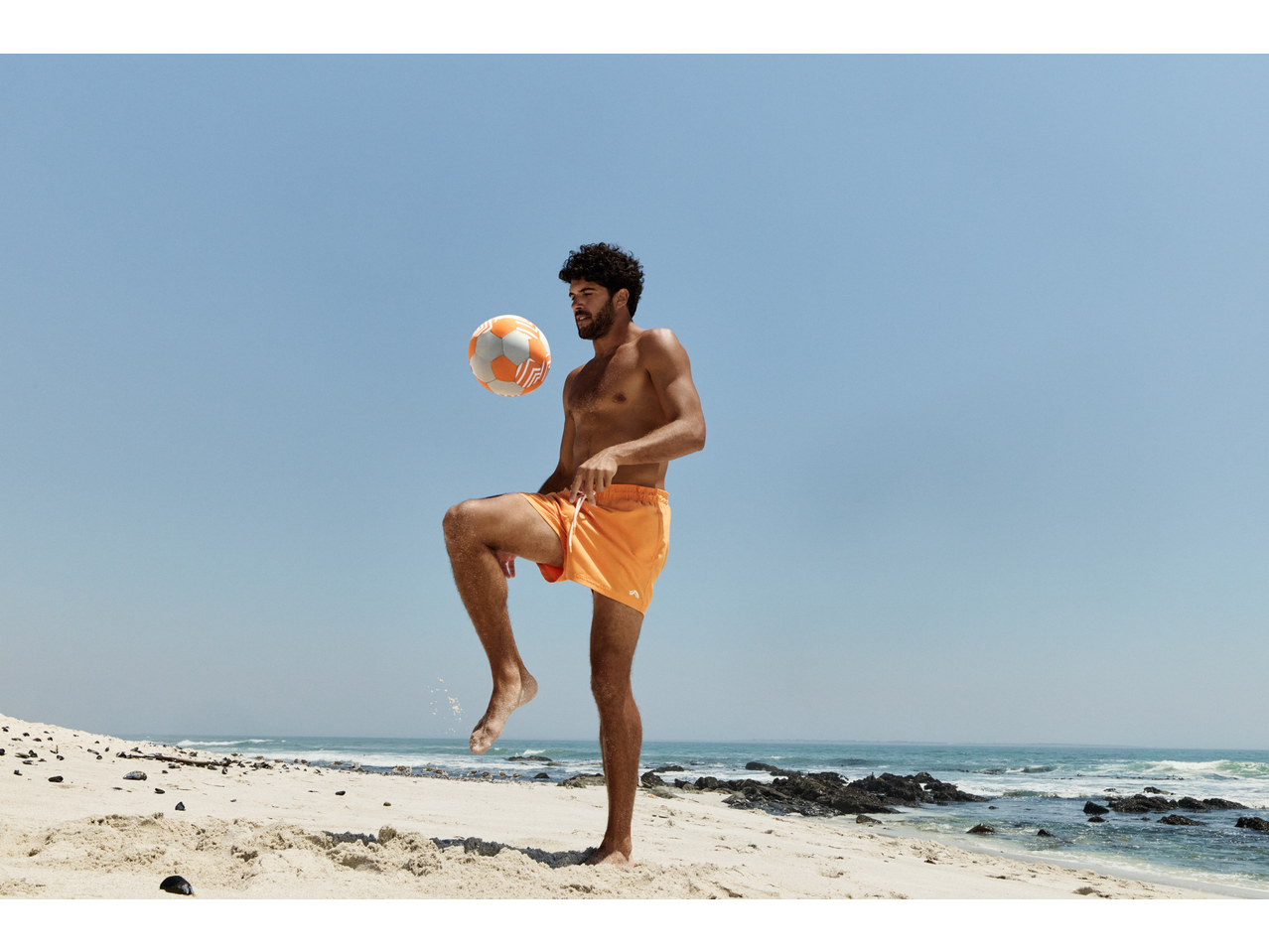 Man in orange CRIVIT shorts playing with a Neoprene Ball on a sunny beach.