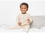 A baby in a beige knitted cardigan and pants sitting on a white blanket.
