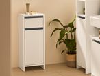 A white narrow bathroom cabinet with two drawers sits beside a potted plant.