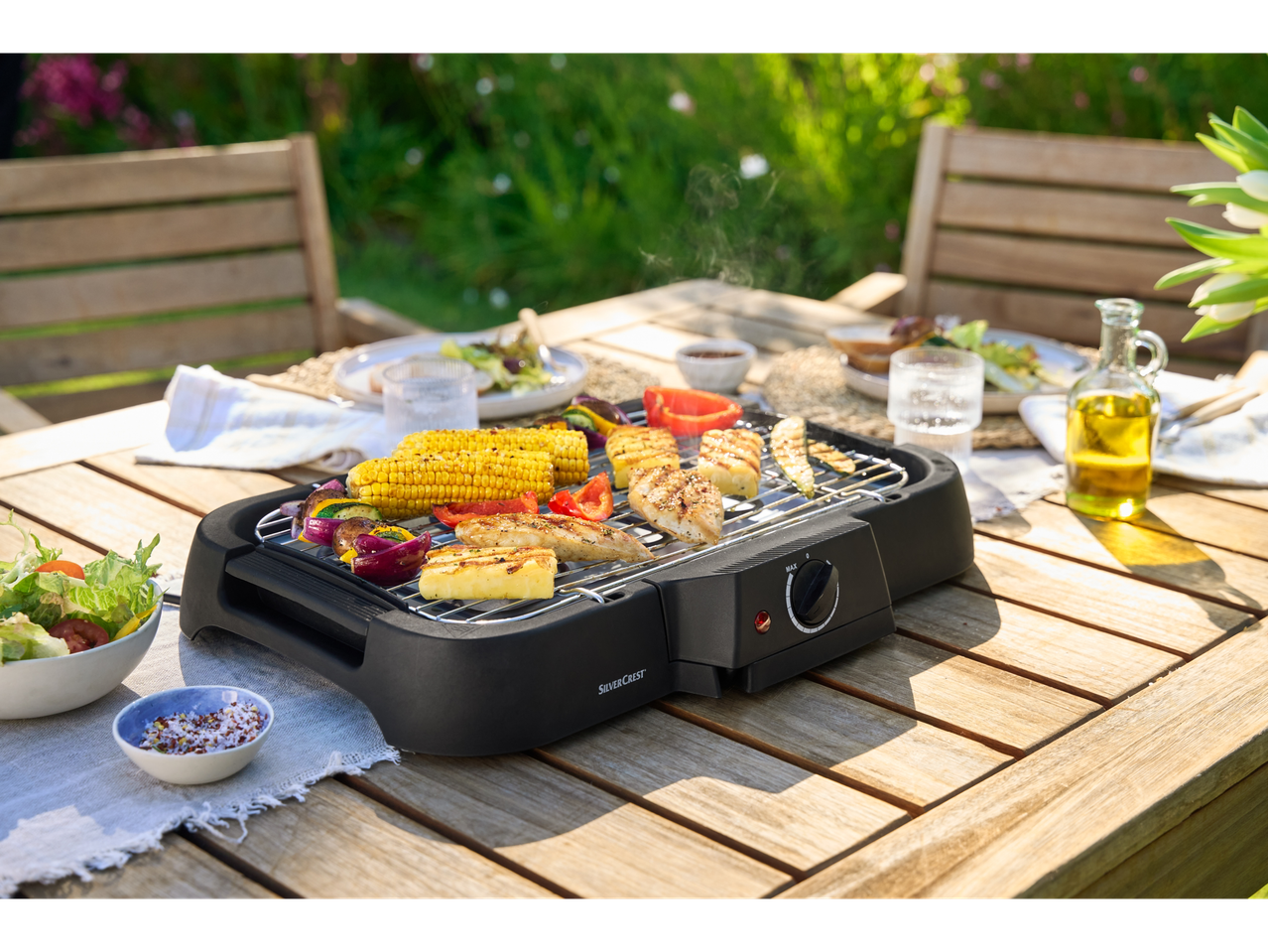Grill elétrica SilverCrest com legumes, frango e abacaxi grelhados em uma mesa de madeira ao ar livre.