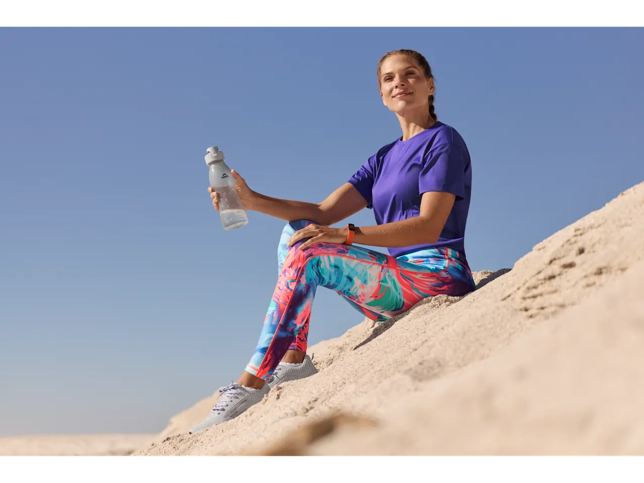 Femeie odihnindu-se pe plajă după antrenament, cu o sticlă de apă CRIVIT și haine sport.