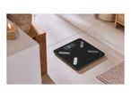 Black smart bathroom scale on a tiled floor, next to a lit candle and wooden furniture.