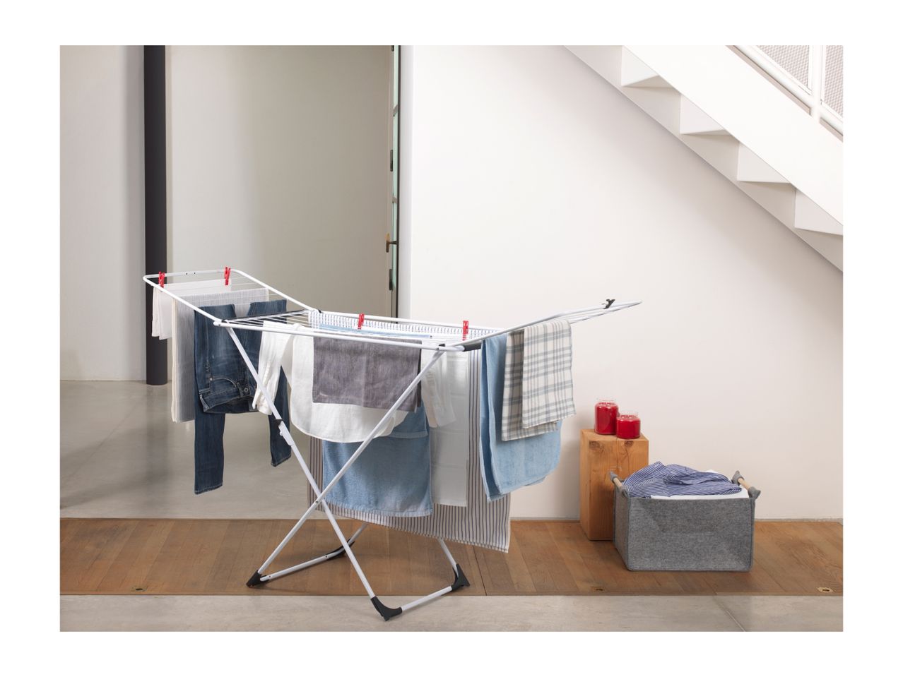 White clothes drying rack with various clothes, next to a grey laundry basket and red candles.