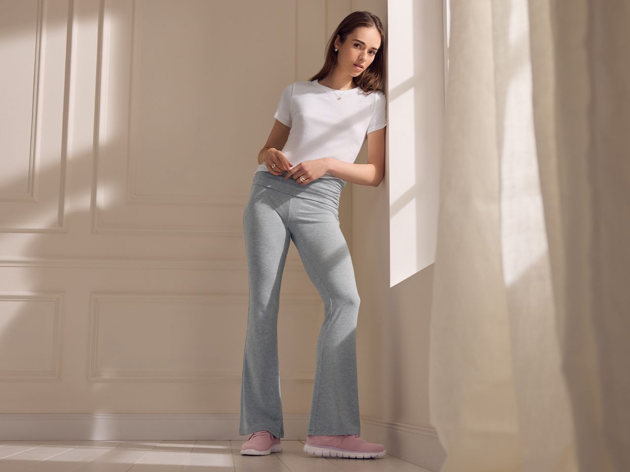 Woman in white t-shirt and grey flared trousers, leaning against a wall.