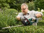 Man in safety glasses and gloves trimming a green hedge with a cordless hedge trimmer.