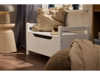 O bancă de depozitare din lemn alb, cu o pernă bej, se află lângă o fereastră, prezentând un stil de decor acasă confortabil.