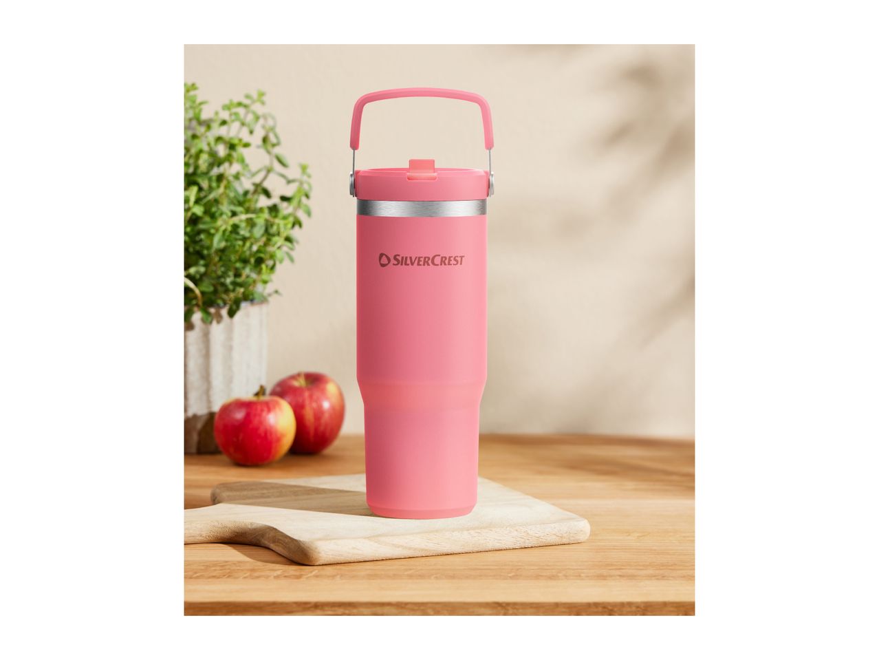 A pink Silvercrest travel mug on a wooden cutting board with apples and a plant.