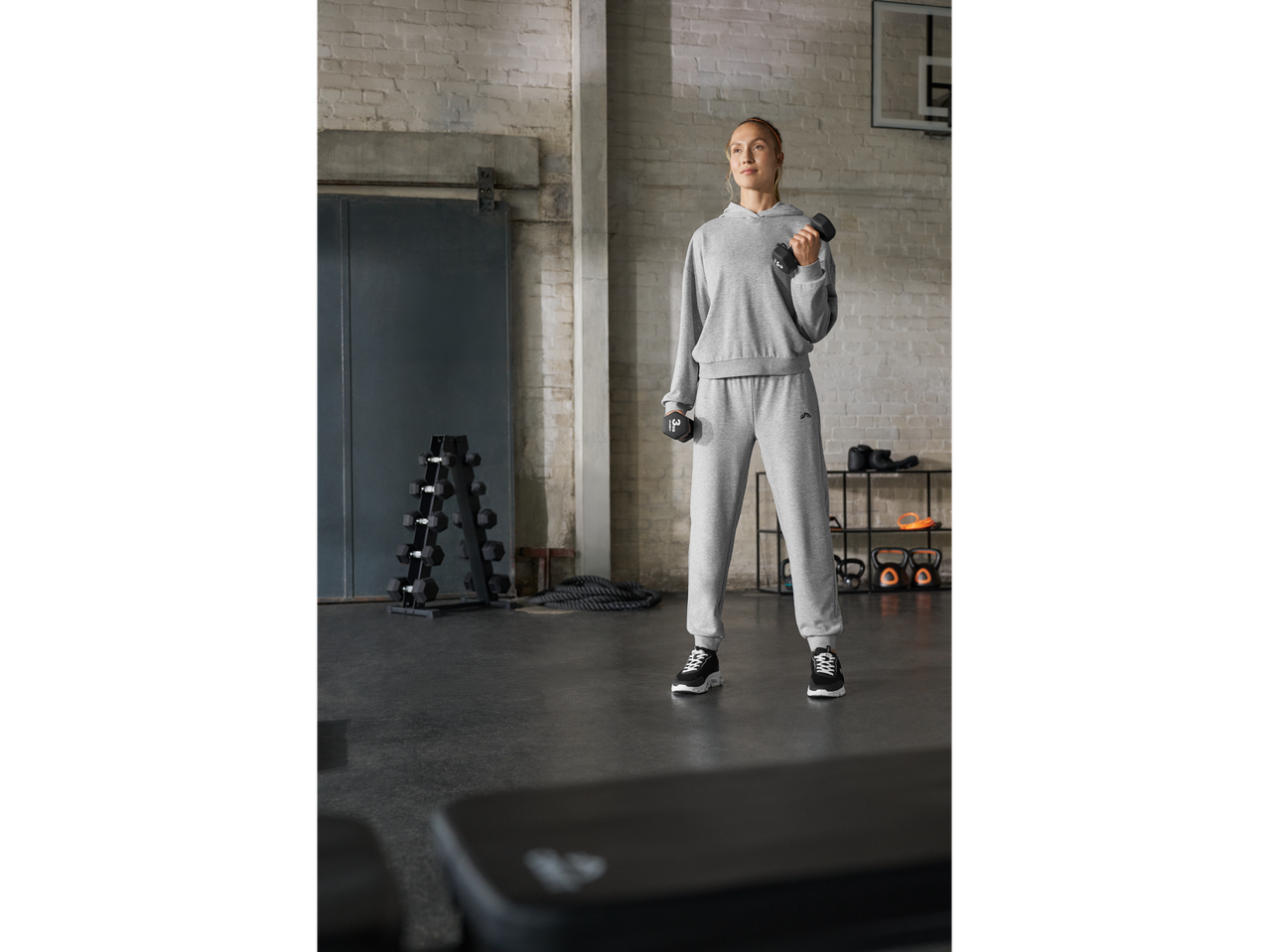A woman in a grey tracksuit holds dumbbells in a gym with a dumbbell rack and kettlebells.