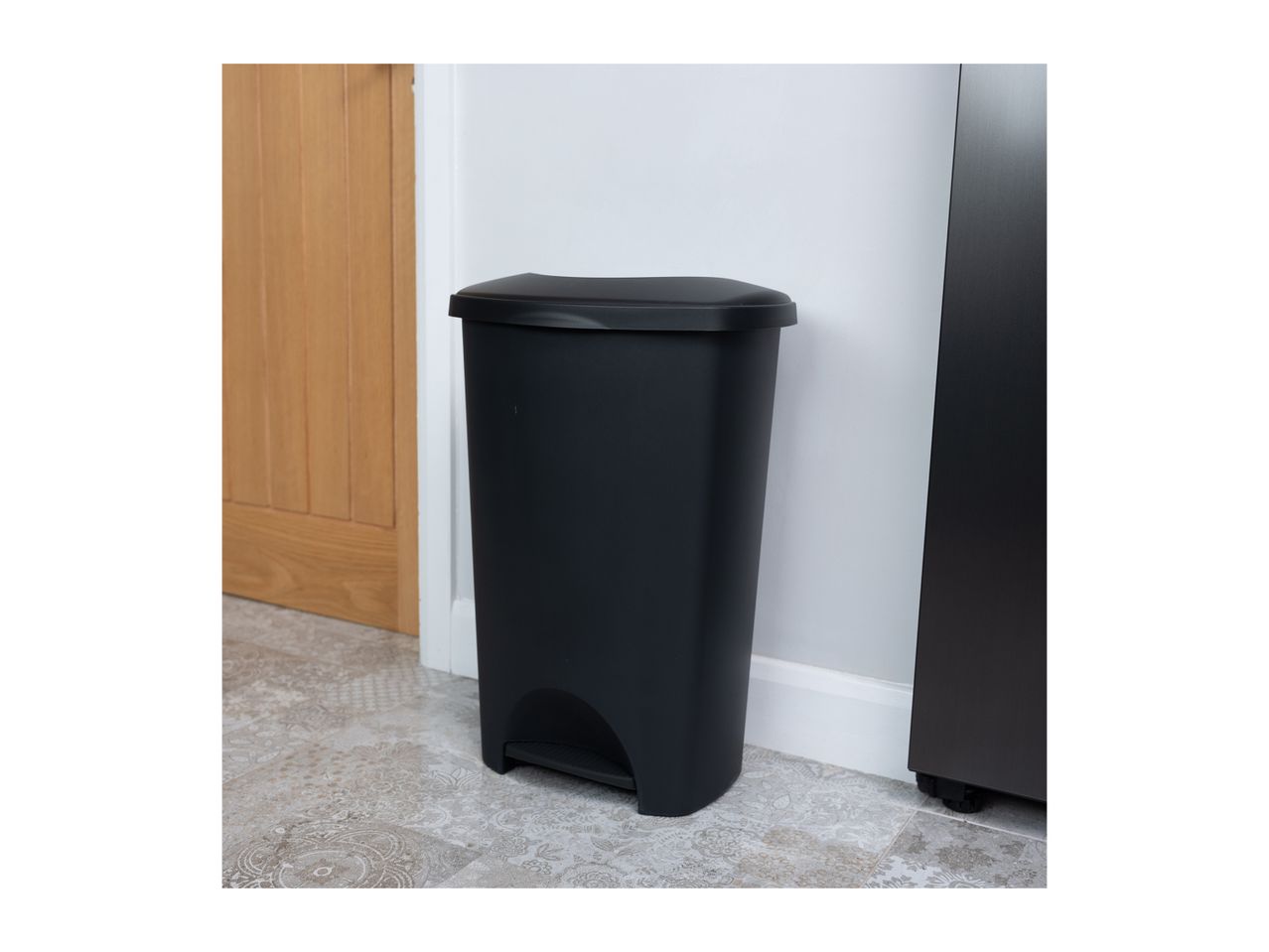 Black pedal bin with a lid, standing on a patterned floor next to a wooden door.