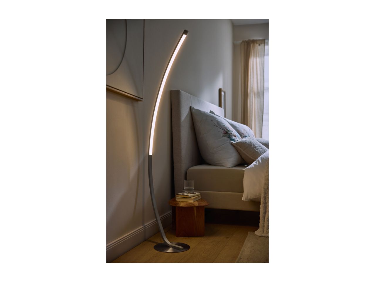 Modern curved LED floor lamp illuminating a cozy bedroom next to a bed.