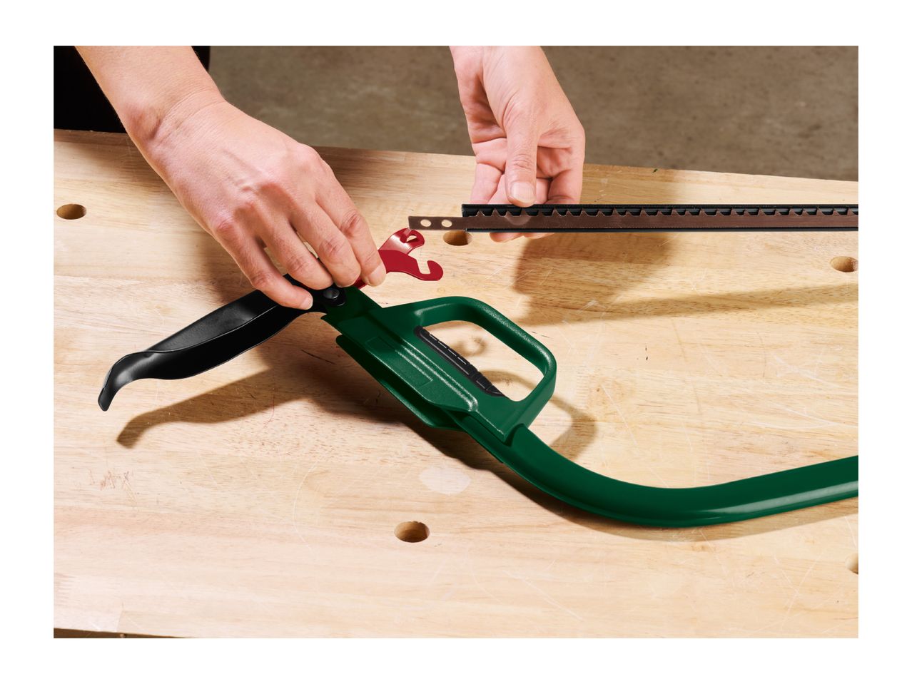 A person attaching a saw blade to a green bow saw frame on a wooden workbench.