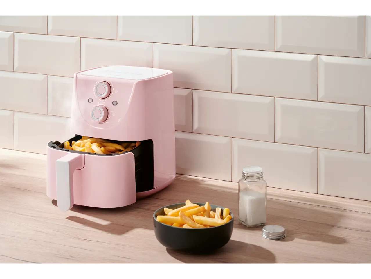 Roze friteza na vazduh sa pomfritom, činijom pomfrita i slanikom na drvenom pultu.