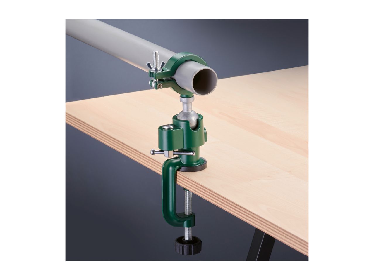 A green table clamp with a pipe holder attached to a light wooden surface, holding a grey pipe.