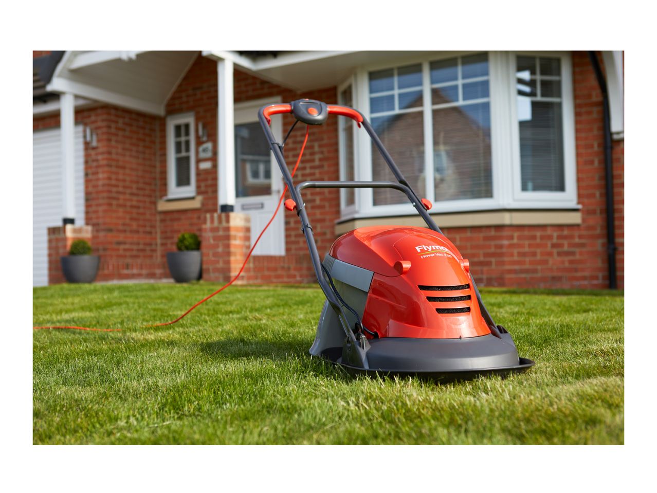An orange and grey hover lawnmower on green grass in front of a brick house.
